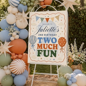 May include: A rectangular birthday sign on a white metal easel, featuring the text 'Welcome to Juliette's 2nd Birthday Two Much Fun'. The sign is decorated with colorful balloons, bunting, and stars, surrounded by a large arch of balloons in blue, green, and orange.