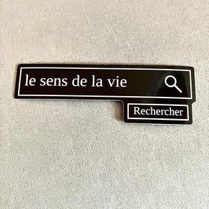 May include: A black sticker with white text that reads "le sens de la vie" and "Rechercher" in a search bar design. The sticker has a white border and a magnifying glass icon. The background is a light gray fabric.