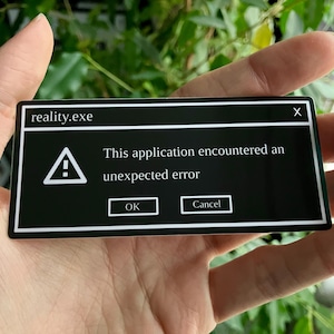 May include: A black sticker with a white border displaying a computer error message. The text reads "reality.exe" at the top, with a warning symbol and the message "This application encountered an unexpected error." Buttons for "OK" and "Cancel" are present.