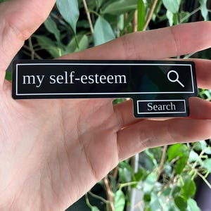 May include: A black sticker with the words "my self-esteem" in white, resembling a search bar. A magnifying glass icon is to the right, and a smaller box below reads "Search". The sticker is held in a hand against a background of green leaves.