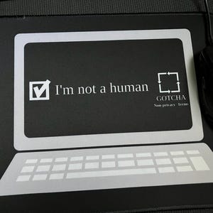 Puede incluir: Ratón y alfombrilla de ratón negros con diseño de portátil. La alfombrilla presenta un contorno de portátil con teclado y pantalla. El texto en la pantalla dice "I'm not a human" con una marca de verificación y un cuadrado con flechas.