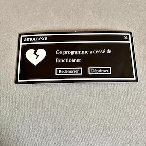 Pode incluir: Um autocolante preto com uma borda branca, apresentando um ícone de coração partido e o texto "amour.exe". O texto "Ce programme a cessé de fonctionner" é exibido, juntamente com os botões "Redémarrer" e "Déprimer".