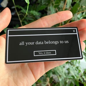 Op de afbeelding: Een zwarte sticker met witte rand, die lijkt op een pop-upvenster van een computer. De tekst "all your data belongs to us" wordt weergegeven, met "Yes, it does" in een kleiner vak eronder. De sticker wordt in een hand gehouden.