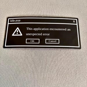 May include: A black sticker with a white border, featuring a computer error message. The text reads "life.exe" at the top, with an exclamation point symbol and the message "This application encountered an unexpected error." Buttons say "OK" and "Cancel."