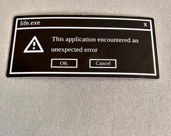May include: A black sticker with a white border, featuring a computer error message. The text reads "life.exe" at the top, with an exclamation point symbol and the message "This application encountered an unexpected error." Buttons say "OK" and "Cancel."