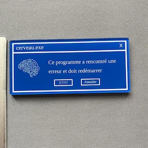 Peut inclure: Un rectangle bleu en acrylique avec du texte blanc et un graphique de cerveau. Le texte indique "cerveau.exe" en haut, avec un message d'erreur simulé en dessous. Le message comprend les boutons "!!??!?" et "Annuler". Une règle est visible sur le côté gauche.