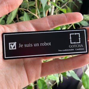 Peut inclure: Une carte rectangulaire noire avec du texte blanc et une coche. Le texte indique "Je suis un robot" et "GOTCHA Non-confidentialité Conditions". La carte est tenue devant un fond de feuilles vertes.