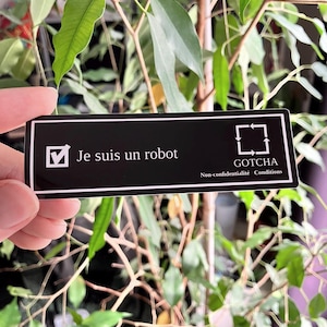 Op de afbeelding: Een zwart rechthoekig bord met witte tekst en een vinkje. De tekst luidt "Je suis un robot" en "GOTCHA Non-confidentialité Conditions". Het bord wordt voor een groene, bladrijke plant gehouden.