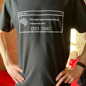Peut inclure: T-shirt noir avec un motif blanc représentant un message d'erreur informatique. Le texte "brain.exe" est en haut, avec un graphique de cerveau et le message "This application encountered an unexpected error." Les boutons indiquent "!!??!?" et "Cancel."