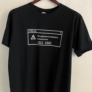 May include: Black t-shirt featuring a white graphic of a computer error message. The text at the top says "reality.exe", with a warning symbol and the message "This application encountered an unexpected error." Below are "OK" and "Cancel" buttons.
