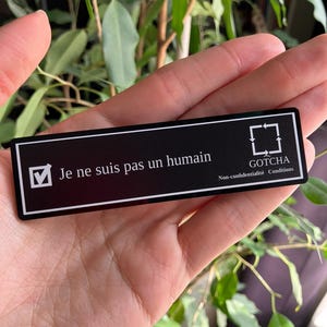 May include: A black rectangular name tag with white text and a checkmark. The text reads "Je ne suis pas un humain" and "GOTCHA" with "Non-confidentialité Conditions" below. The tag is held in a hand with a green plant in the background.
