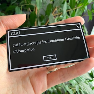 Peut inclure: Un autocollant rectangulaire noir avec du texte blanc et un bord blanc. Le texte indique "CGU" en haut, suivi de texte en français et d'un bouton "Non". L'autocollant est tenu dans une main sur un fond de feuilles vertes.