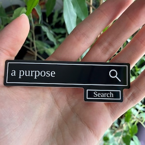 Può includere: Un adesivo rettangolare nero con le parole "a purpose" in bianco, che ricorda una barra di ricerca. Un'icona a forma di lente d'ingrandimento è a destra. Sotto, un rettangolo più piccolo dice "Search".