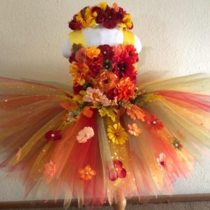 May include: A tutu dress featuring a bodice covered in red, orange, and yellow artificial flowers and leaves. The skirt is made of layered tulle in shades of red, orange, and yellow with scattered floral accents. A matching floral headband is included.
