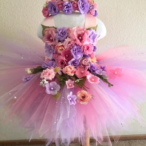 May include: A tutu dress featuring a bodice covered in artificial pink and purple roses, accented with small white pearls. The skirt is made of layered pink and purple tulle with scattered sparkles. A matching floral headband sits on top.