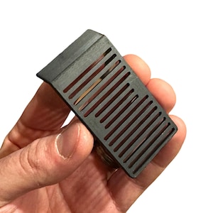 May include: A dark gray, rectangular object with a series of parallel slots. The object has a slightly angled top and is held in a person's hand against a white background. The object appears to be made of a solid material.