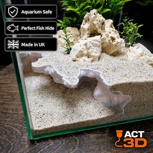 May include: An aquarium cave insert featuring a white, textured, multi-level structure embedded in light beige sand. The scene includes porous, off-white rock formations and green plastic plants. Text overlays state: Aquarium Safe, Perfect Fish Hide, Made in UK.