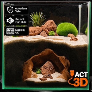 Könnte beinhalten: Ein transparentes Glasaquarium mit einer Querschnittsansicht eines sandigen Bodengrunds und einer unterirdischen Höhlenstruktur. Das Innere enthält braune, strukturierte Steine, eine leuchtend grüne Mooskugel und Pflanzen. Der Text lautet Aquarium Safe, Perfect Fish Hide, Made in UK und ACT 3D.