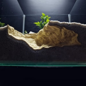 May include: A beige and grey aquarium decoration with a textured, layered design resembling a rock formation. The foreground features a light beige, cave-like structure, while the background is a darker grey, sandy material. Green plants are visible in the background.