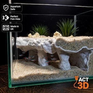 Può includere: Una struttura a grotta per acquario bianca con una superficie superiore sabbiosa e piante artificiali verdi, posizionata all'interno di una vasca in vetro. Piccoli pesci neon nuotano sotto i pilastri bianchi. Il testo include Aquarium Safe, Perfect Fish Hide, Made in UK e ACT 3D.