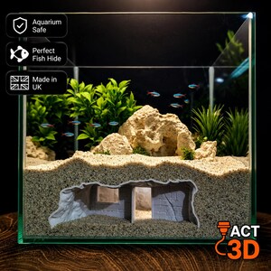 May include: A grey, stone-textured aquarium hideout with a rectangular doorway and window, partially buried in light-coloured sand. The structure features a brick-patterned wall section and is placed inside a glass tank with green plants and rocks.