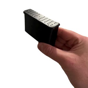 May include: A black rectangular object with small holes on the top, held in a hand against a white backdrop. The object is made of a solid material. The holes are evenly spaced across the surface.