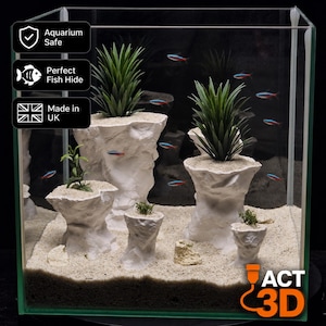 May include: A glass aquarium containing five white, rock-like structures of varying heights, each topped with green artificial plants. Small neon tetra fish swim around the structures, which are set on a light-coloured sand substrate. Text overlays include Aquarium Safe, Perfect Fish Hide, Made in UK, and the logo ACT 3D.