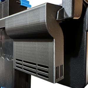 May include: A close-up of a black plastic component featuring horizontal and vertical vents. The component has a curved design and is part of a larger structure. A blue plastic piece is visible at the top.