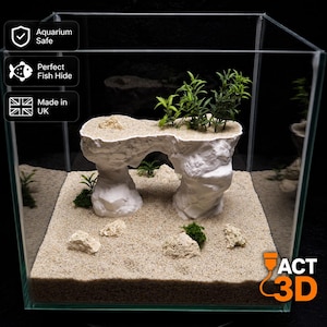 May include: A white rock-like aquarium ornament with an archway, topped with green plastic plants, placed on light beige sand inside a clear glass tank. Text on the image includes Aquarium Safe, Perfect Fish Hide, Made in UK, and ACT 3D.