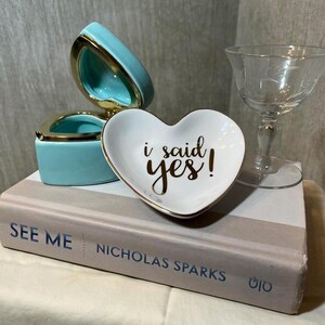 May include: A heart-shaped trinket dish with the words "i said yes!" in gold lettering, a turquoise ring box, and a champagne glass are arranged on a book. The ring box is open, revealing a gold interior.