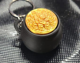 May include: A black, spherical, pot-shaped keychain featuring a gold-colored insert filled with a textured, circular arrangement of stacked coin-like shapes. The object includes a standard metal split ring attached to the top for keys.