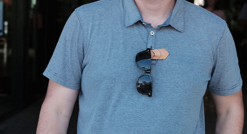 Op de afbeelding: Een blauw poloshirt met een leren zonnebrilhouder die aan de voorkant is bevestigd. De zonnebril is zwart en heeft reflecterende glazen.