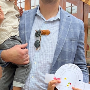 Op de afbeelding: Een man draagt een blauw geruit blazer en een wit overhemd met knopen en een klein patroon. Hij houdt een zonnebril vast die aan een bruin leren bandje is bevestigt. De zonnebril hangt aan het bandje op zijn borst. Hij houdt ook een wit vel papier vast met een rood hart en gele sterren erop. Het papier heeft de tekst "Jezus wil mijn vriend voor altijd zijn" erop.