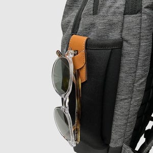 May include: A gray and black backpack with a brown leather strap and a clear glasses holder. The glasses holder is attached to the strap and is designed to hold sunglasses or reading glasses.