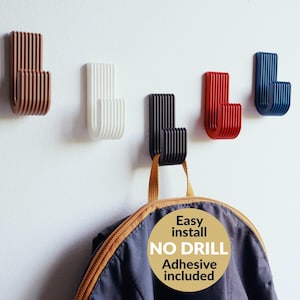 May include: Five wall hooks in various colors: brown, white, black, red, and blue. Each hook has a curved, ribbed design. A jacket hangs from the black hook. The image includes text that says "Easy install, NO DRILL, Adhesive included."