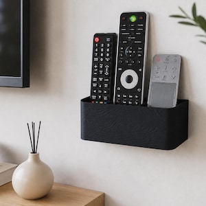 Peut inclure: Un support mural noir pour télécommandes contenant trois télécommandes. Les télécommandes sont noires et argentées. Le support est fixé sur un mur blanc près d'une télévision.