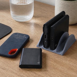 Puede incluir: Una configuración de escritorio con una computadora portátil, un ratón y varios accesorios tecnológicos. Una funda de teléfono azul oscuro con un acento naranja, un pequeño banco de energía negro y dos dispositivos negros en un soporte gris están dispuestos sobre una superficie de madera.