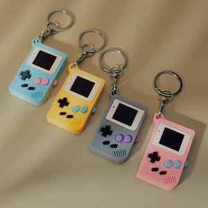 以下が含まれることがあります： レトロな携帯ゲーム機型のカラフルなキーホルダー4個セット。キーホルダーはパステルブルー、イエロー、グレー、ピンクです。それぞれ黒い画面、十字キー、カラーボタンが付いています。キーホルダーはシルバーのキーリングに付いています。