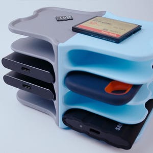 Peut inclure: Un organisateur de rangement à plusieurs niveaux en bleu clair et gris, conçu pour contenir divers accessoires technologiques. Il comporte plusieurs emplacements pour disques durs externes, cartes SD et autres petits appareils. L'organisateur est en plastique et a un design moderne.