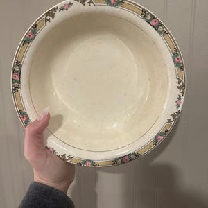 Puede incluir: Cuenco de cerámica redondo de color crema con un borde floral decorativo en rosa, verde y dorado. El cuenco está sostenido por una persona. El borde tiene un ribete dorado.