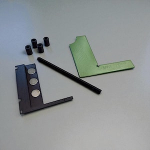 May include: An assortment of black and green metal components, including a right-angle piece, a long rod, and several small cylinders. The components are arranged on a gray surface, suggesting a mechanical or engineering theme.