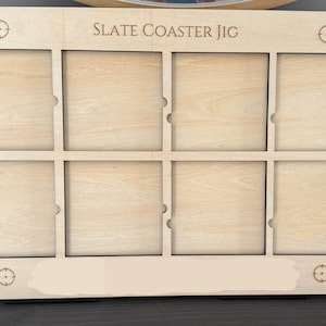 May include: A light-colored wooden slate coaster jig with eight square compartments. The text "SLATE COASTER JIG" is engraved across the top. Each corner has a target symbol.
