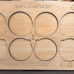 May include: A wooden slate coaster jig with eight circular cutouts. The text "SLATE COASTER JIG" is engraved across the top. The jig has a light wood grain finish and features decorative target symbols.
