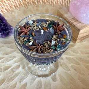 May include: A dark purple candle in a clear glass goblet, decorated with star anise, small crystals, and dried herbs. The candle sits on a decorative surface, with a purple amethyst and a pink crystal in the background.