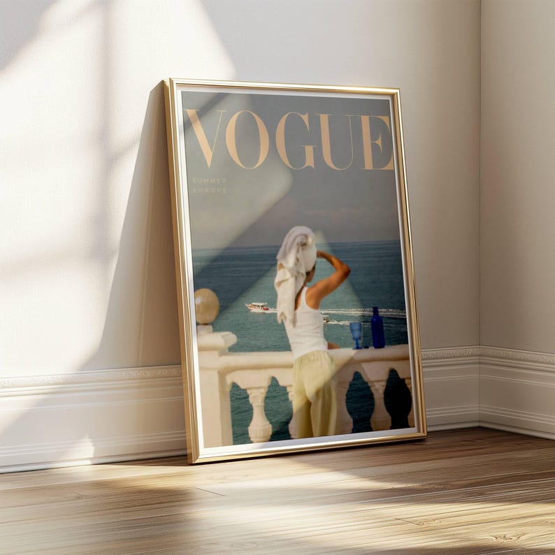 Pode incluir: Uma capa emoldurada da revista Vogue com uma mulher numa varanda com vista para o mar. A capa da revista tem a palavra "VOGUE" em letras douradas. A mulher est&aacute; vestida com um top branco e cal&ccedil;as amarelas. Uma garrafa e um copo azuis est&atilde;o na varanda.