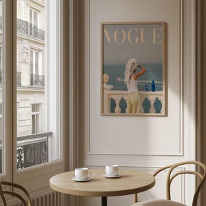 Pode incluir: Uma impress&atilde;o emoldurada da capa da revista "Vogue". A imagem apresenta uma pessoa numa varanda com vista para o mar. A pessoa est&aacute; vestida com um top branco e cal&ccedil;as amarelas. Uma garrafa azul est&aacute; na varanda. A impress&atilde;o est&aacute; numa moldura de madeira clara.