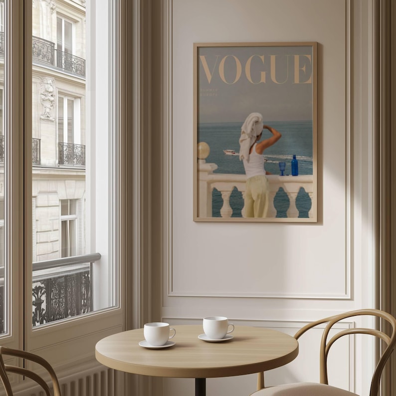 Pode incluir: Uma impress&atilde;o emoldurada da capa da revista "Vogue". A imagem apresenta uma pessoa numa varanda com vista para o mar. A pessoa est&aacute; vestida com um top branco e cal&ccedil;as amarelas. Uma garrafa azul est&aacute; na varanda. A impress&atilde;o est&aacute; numa moldura de madeira clara.