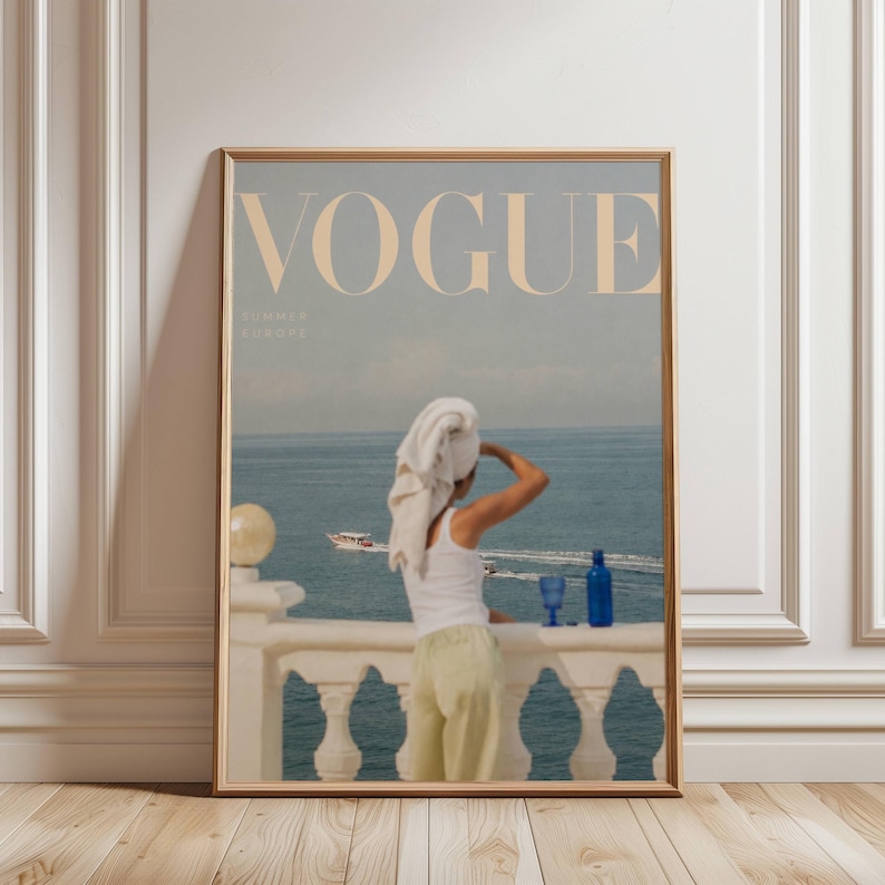 Pode incluir: Uma impress&atilde;o emoldurada com uma capa de revista Vogue com o texto "VOGUE SUMMER EUROPE". A imagem mostra uma pessoa em uma varanda com vista para o mar, usando uma toalha na cabe&ccedil;a. Copos azuis e uma garrafa na varanda.