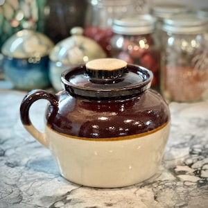 May include: A two-toned ceramic crock with a lid and handle. The upper portion is a glossy dark brown, while the lower section is a creamy beige. A wooden knob sits atop the lid. Other jars are visible in the background.