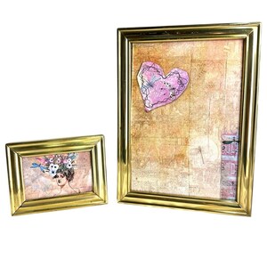 May include: Two gold-framed art pieces. One frame contains a portrait of a woman with a floral headdress. The other frame holds a pink heart-shaped design with a dragonfly illustration. Both frames have a rectangular shape.